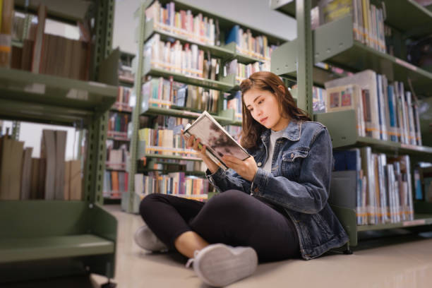 Asian female student sitting on floor in the library, Open and learning textbook from bookshelf Asian female student sitting on floor in the library, Open and learning textbook from bookshelf in the International College/University Library. female high school student stock pictures, royalty-free photos & images