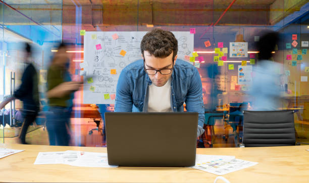 mann, der in einem kreativen büro mit seinem computer arbeitet und menschen, die sich im hintergrund bewegen - innovation stock-fotos und bilder