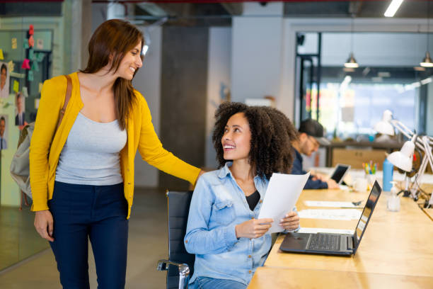 Business woman arriving to the office and greeting a coworker at her desk Latin American business woman arriving to the office and greeting a coworker at her desk - lifestyle at the place of work office leave stock pictures, royalty-free photos & images