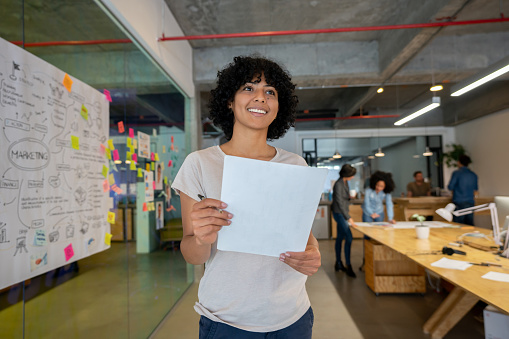 Happy African American woman working at a creative office and thinking of ideas for a project while holding a document