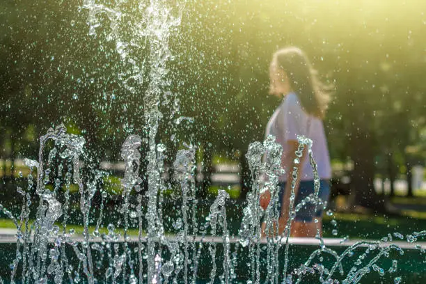 Photo of Cityscape with gush water of fountain. Silhouette of young woman walking behind splashes of fountain on hot summer day.