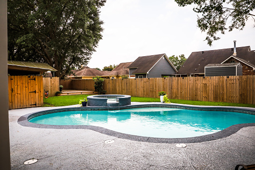 Una gran piscina de estilo gris gris de forma libre con agua azul turquesa en un patio trasero cercado en un vecindario de suburbios. photo