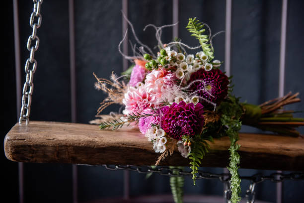 On a wooden board swing lies a wedding bouquet suspended on iron chains in a cage in a dark loft interior. Unusual flowers for the holiday: green fern, chrysanthemums, dahlias, pampas grass, dried flowers en un columpio de tabla de madera se encuentra el ramo de la boda suspendido en cadenas de hierro en una jaula en el interior oscuro loft. flores inusuales para las vacaciones: helecho verde, crisantemos, dalias, hierba de las pampas, flores secas