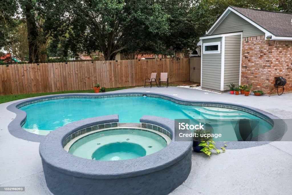 A large free form gray grey accent swimming pool with turquoise blue water in a fenced in backyard in a suburb neighborhood. A large free form gray grey accent swimming pool and hot tub Hot Tub, turquoise blue swim water in a fenced in backyard in a suburb neighborhood. Back Yard Stock Photo