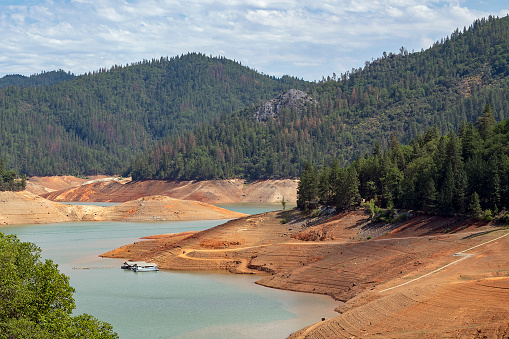 Low water level at Shasta Lake, California, USA due to multi-year drought. The lake is 100 feet/33 meters below full capacity. Paved boat ramp in lower right no longer reaches water. Dirt boat ramp provides temporary access. Houseboats moored near swimming platform who's underwater anchor, which should be vertically below platform, lies exposed horizontally on dry shore. Shasta Lake provides recreation as well as drinking water and irrigation water to locations as far away as Los Angeles 500 miles/800km away. June 21, 2021.