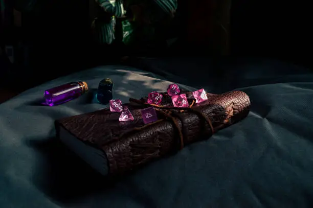 Image of pink transparent gaming dice on a leather-bound notebook partially lit by the sun. In the background two potions in glass stopper bottles