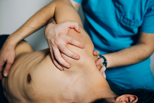 Close up of unrecognizable handsome young man lying on side on the bed while therapist adjusting his shoulder holding his arm