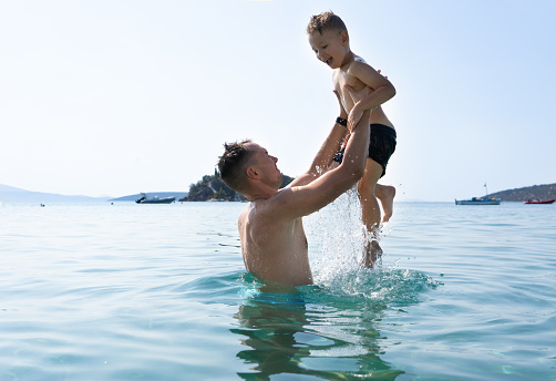 Dad and son are playing in the sea. The father tosses the son up. Greek islands, water splashes, vacations, blue sky and sunny day. High quality photo