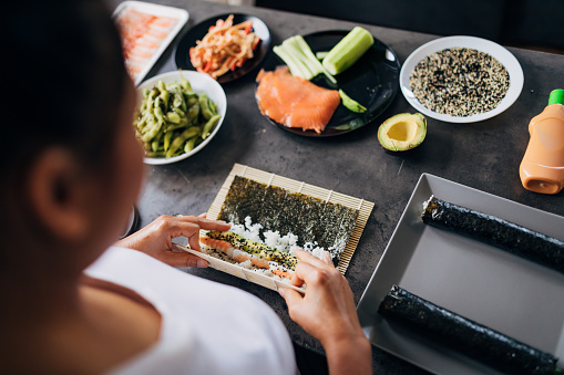 In her kitchen woman Thai ethnicity, making traditional sushi rolls, with fresh and raw vegetables and sea food, while using bamboo mat to roll it up