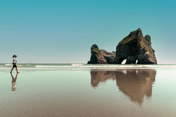 mujer caminando por la playa de wharariki y formaciones rocosas increíbles reflejadas en la arena. nueva zelanda - golden bay fotografías e imágenes de stock