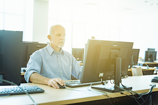 High key image of a businessman at work in an open plan office with multiple workstations engrossed in his desktop computer backlit with bright sun flare through a large window