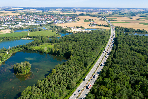 Heavy car and truck traffic on rural highway among green forests and lakes, aerial view.