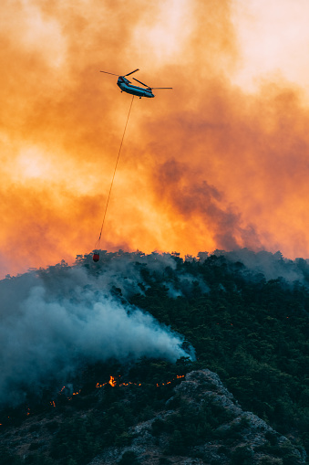 ronda, Spain – June 11, 2022: ronda,malaga,spain june 11, 2022 firefighting helicopter in mid-flight dropping water with the sky red from the flames of the fire.