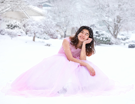 Biracial teen girl in long formal pink gown sitting outdoors in beautiful snowfall