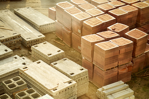 Top view of stacks of cement floor slabs and rectangular red perforated bricks packed in cellophane placed on the wooden pallets lying on the sunlit sandy ground