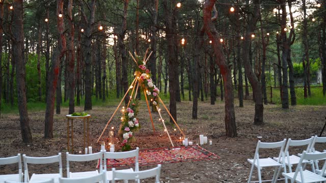 Festive string lights illumination on boho tipi arch decor on outdoor wedding ceremony venue in pine forest at night. Vintage string lights bulb garlands shining above chairs at summer rural wedding