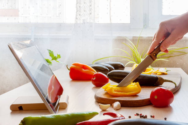Woman slicing pepper watching online culinary class her tablet, cooking vegan food with fresh vegetables. stock photo