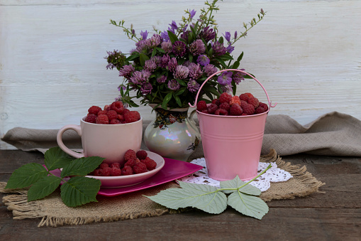There is a bouquet of clovers on the table, raspberry leaves and a bucket and a cup of raspberries.