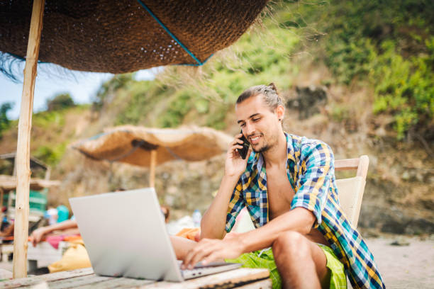 Working could be pleasure. Young man working on his laptop on the beach and talking on the phone. Leisure activities / Remote working concept. digital nomad stock pictures, royalty-free photos & images