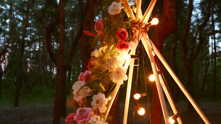 Bohemian tipi wooden arch decorated with burning candles, roses and pampass grass, wrapped in fairy lights illumination on outdoor wedding ceremony venue in pine forest at night. Bulbs garland shines