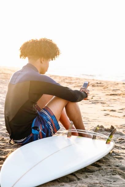 istock Afro surfer man consulting application on smartphone with surfboard on beach at summer sunrise 1332273086