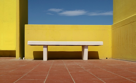 White concrete bench with terracotta tiles on the pavement.   Bright yellow concrete wall and blue sky with cloud on behind. Background for copy space