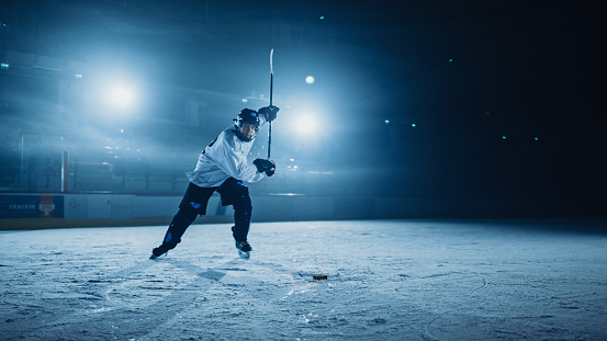 Ice Hockey Rink Arena: Professional Player Shooting, Hitting, Stricking the Puck with Hockey Sticks. Athlete Scoring a Goal. Dramatic Wide Shot, Cinematic Lighting.