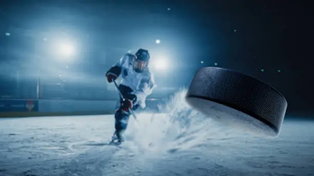 Photo of Ice Hockey Rink Arena: Professional Player Shooting the Puck with Hockey Stick. Focus on 3D Flying Puck with Blur Motion Effect. Dramatic Wide Shot, Cinematic Lighting.