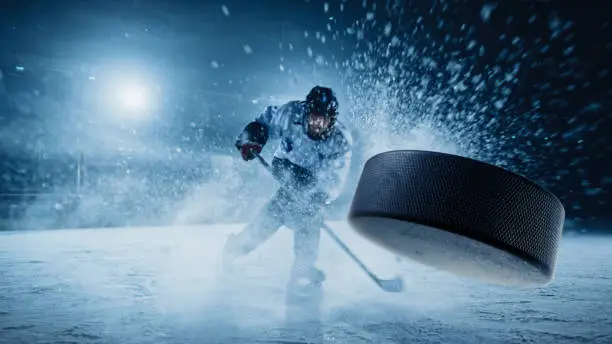 Photo of Ice Hockey Rink Arena: Professional Player Shooting the Puck with Hockey Stick. Focus on 3D Flying Puck with Blur Motion Effect. Dramatic Wide Shot, Cinematic Lighting.