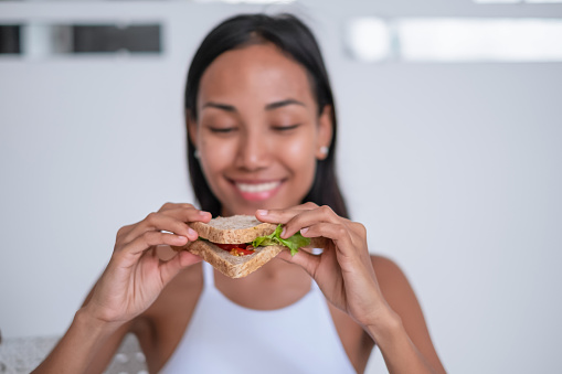 Front view shot of a cheerful Indonesian woman eating a vegetarian sandwich made of various vegetables and whole-grain loaf.