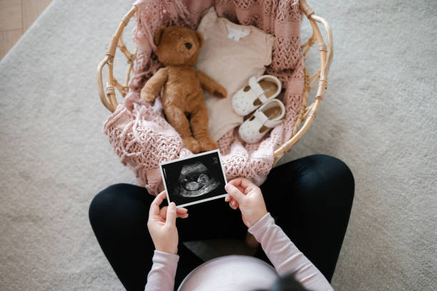 Overhead view of Asian pregnant woman holding an ultrasound scan photo of her baby, with a moses basket filling with baby clothing, baby shoes and soft toy teddy bear in front of her. Mother-to-be. Expecting a new life. Preparing for the new born concept stock photo