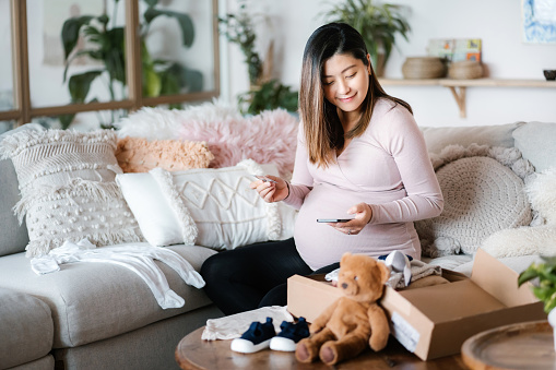 Smiling Asian pregnant woman shopping online with smartphone and making mobile payment with credit card in the living room. A delivery package of baby clothing and toys on coffee table. Time to get some baby essentials for her unborn baby!
