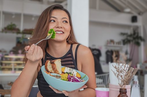 Candid shot of a cheerful young Chinese woman eating a healthy vegan salad with sourdough bread in a cafe. She's holding a fork with a piece of broccoli on it.