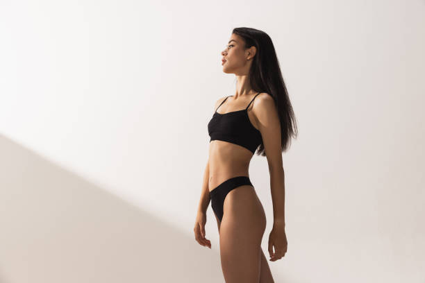 Studio image of young beautiful woman with slim figure in black underwear posing over white studio background Side view portrait of one young beautiful tanned woman with slim figure in black underwear standing on white studio background. Concept of female beauty, flexibility, tenderness bikini stock pictures, royalty-free photos & images