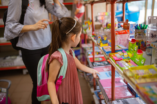 Charming preschool/school girl, with her caring mother buying her school supplies in the market-retail place, ready for her first day at school