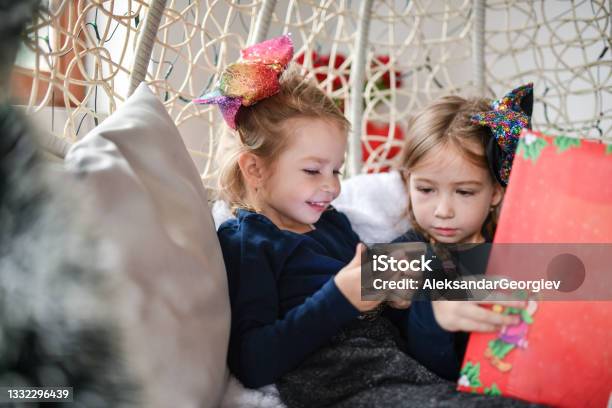 Cute Child Sisters Sitting In Home Swing And Comparing Smartphone To Graphic Novel Stock Photo - Download Image Now