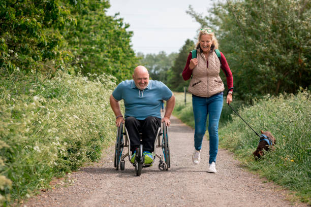 Using My Wheelchair on a Dog Walk A caucasian, mature man with paraplegia and his friend, a mature caucasian woman, on a day out in a nature reserve with her dog on a sunny summer's day. The man is using his modified wheelchair while his friend is walking with her dog on a leash. They are both wearing casual clothing and are having a positive day out together. dog disruptagingcollection stock pictures, royalty-free photos & images