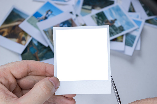 A person holds a polaroid photo in his hands against the background of other photos. Photo in hand close-up. A person looks at a photo from a polaroid.