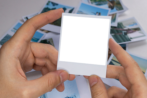 A person holds a polaroid photo in his hands against the background of other photos. Photo in hand close-up. A person looks at a photo from a polaroid.