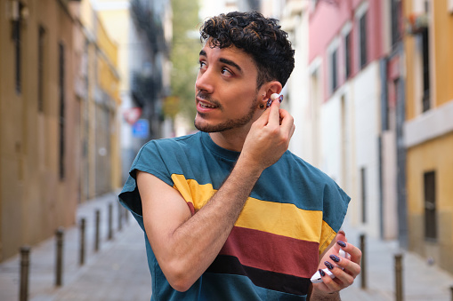 Young caucasian man with long false nails putting on his wireless earbuds at street.