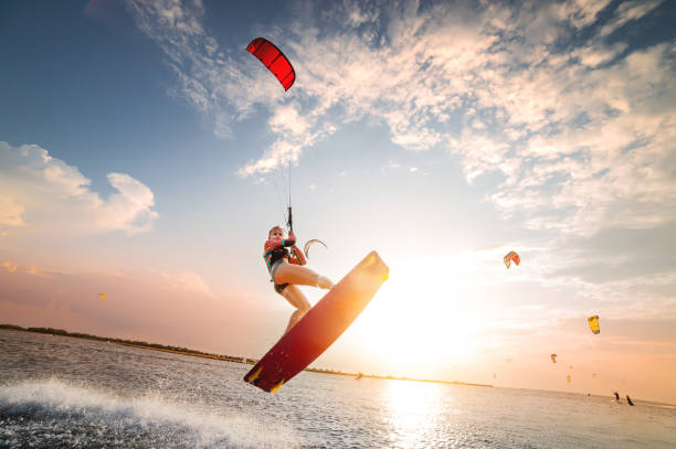 Sports shot of a young caucasian woman in a wetsuit doing a trick in the air against the backdrop of a sunset in the sea. Kitesurring girl athlete flies on a kite with a board stock photo
