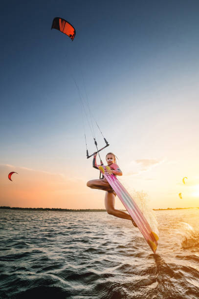 広角。ウェットスーツを着た若い白人女性は、海の夕日を背景に水上を滑るトリックを作ります。ボードで凧の上にカイトサリング女の子アスリートダークサイドトリック - women sensuality surfing water sport ストックフォトと画像