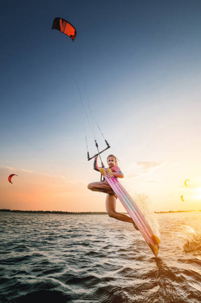 Wide angle. young caucasian woman in a wetsuit makes a trick sliding on the water against the backdrop of a sunset in the sea. Kitesurring girl athlete dark side trick on a kite with a board stock photo