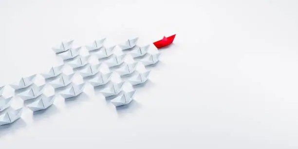 Arrow shaped group of white paper boats on a white background with a single leader boat ahead