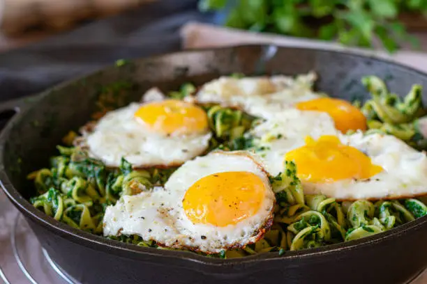 Homemade vegetarian pasta dish with creamy spinach and fried eggs, sunny side up. Served in a rustic cast iron pan on a table. Isolated view