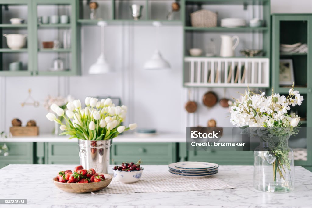 Kitchen countertop with flowers in bucket and jar Kitchen island with marble surface, small bowl of cherries with bigger wooden bowl filled with strawberry, flowers in bucket and glass jar, green vintage cupboards full of different devices Home Interior Stock Photo
