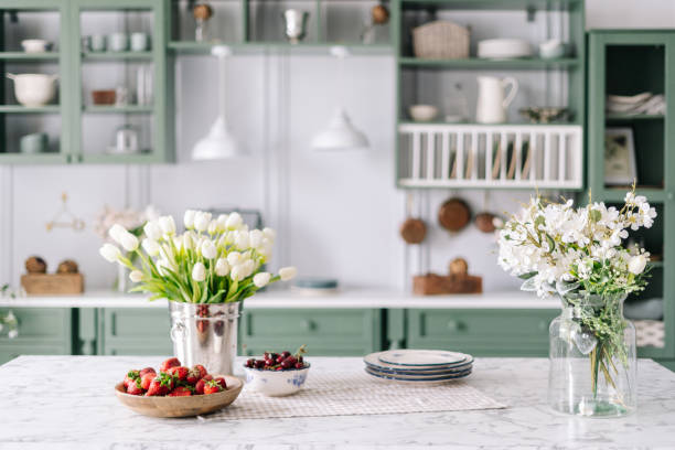Kitchen countertop with flowers in bucket and jar Kitchen island with marble surface, small bowl of cherries with bigger wooden bowl filled with strawberry, flowers in bucket and glass jar, green vintage cupboards full of different devices hygge photos stock pictures, royalty-free photos & images