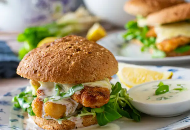 homemade fresh cooked fish burger with redfish fillet, cheese, delicious sauce served on a whole grain bun on a table. Closeup and front view with blurred background