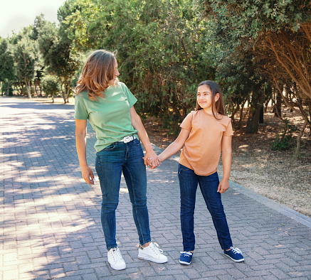 Mother and daughter walking in the park. A middle-aged woman and kid girl holding each other hands, making eye contact and wearing green and orange t-shirts