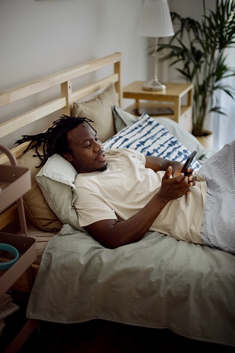 The young man relaxes at home while lying in bed.  Black man on a video call . Morning routine with phone and technology